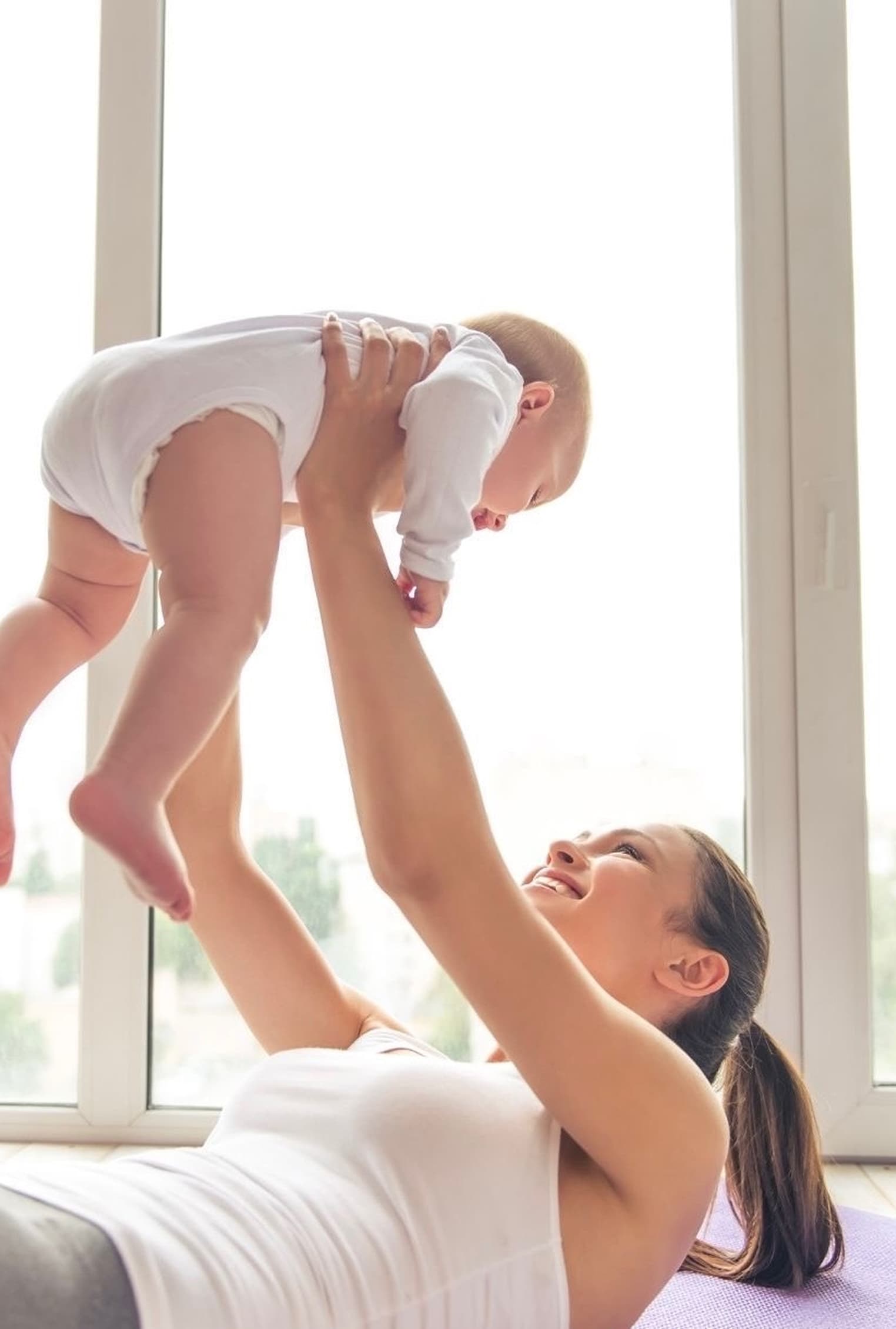 A mother smiling at her baby while laid on a yoga mat.